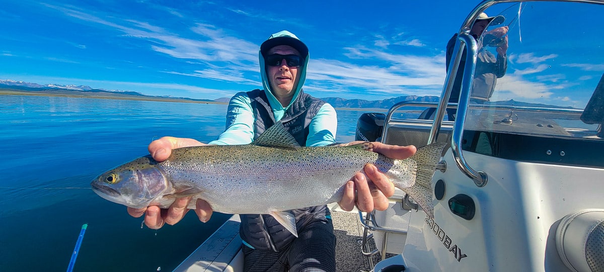 A smiling fly fisherman on a lake in a boat holding a large rainbow trout.