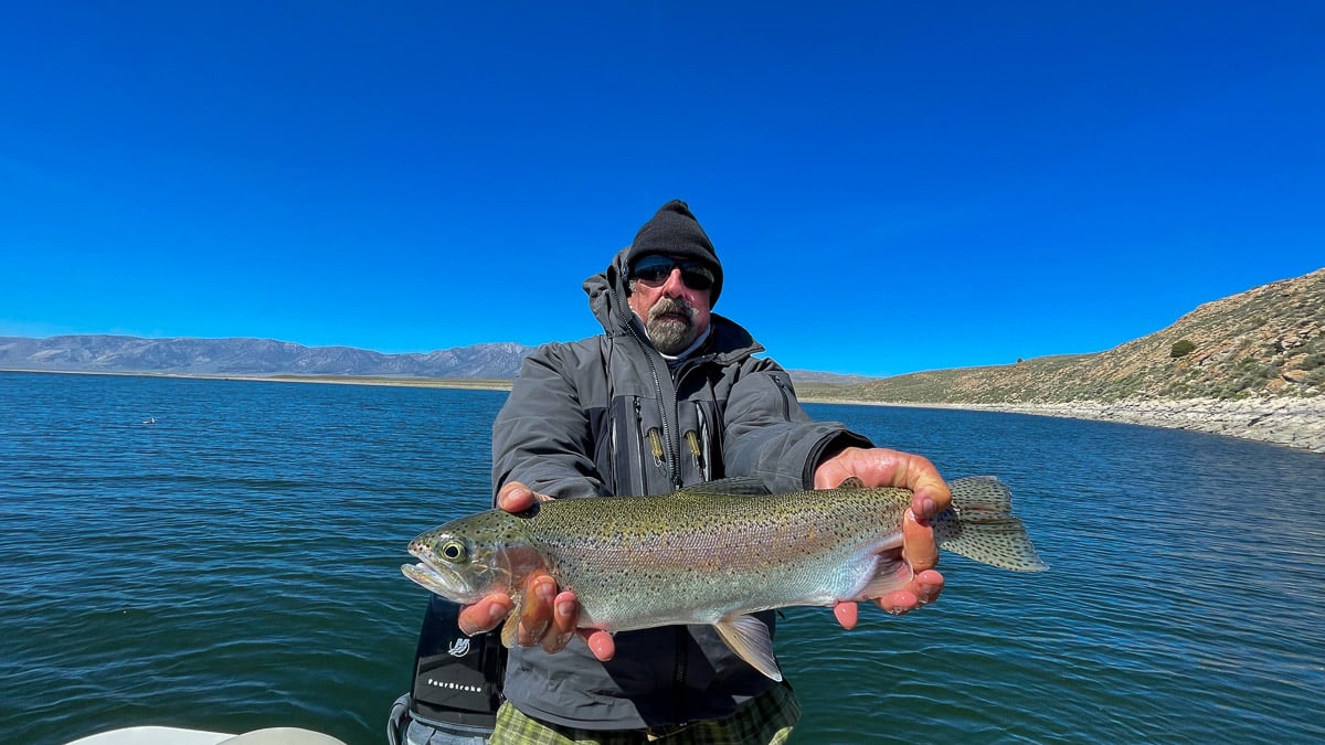 A smiling fly fisherman on a lake in a boat holding a large rainbow trout.