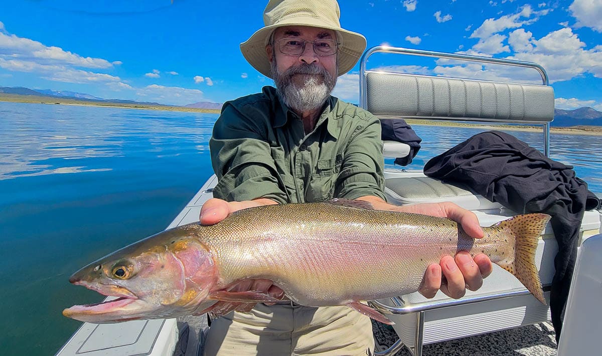 A smiling fly fisherman on a lake in a boat holding a large cutthroat trout.