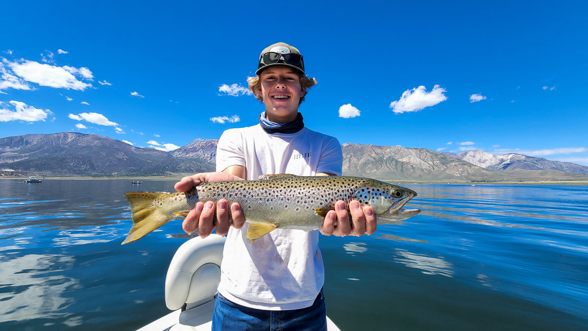 A smiling angler holding a large rainbow trout in a boat on a lake.