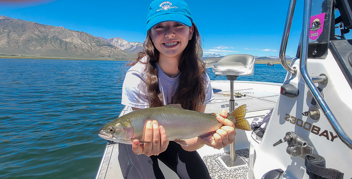 A smiling female fly fisherman and a young girl wearing pink on a lake in a boat holding a large rainbow trout.