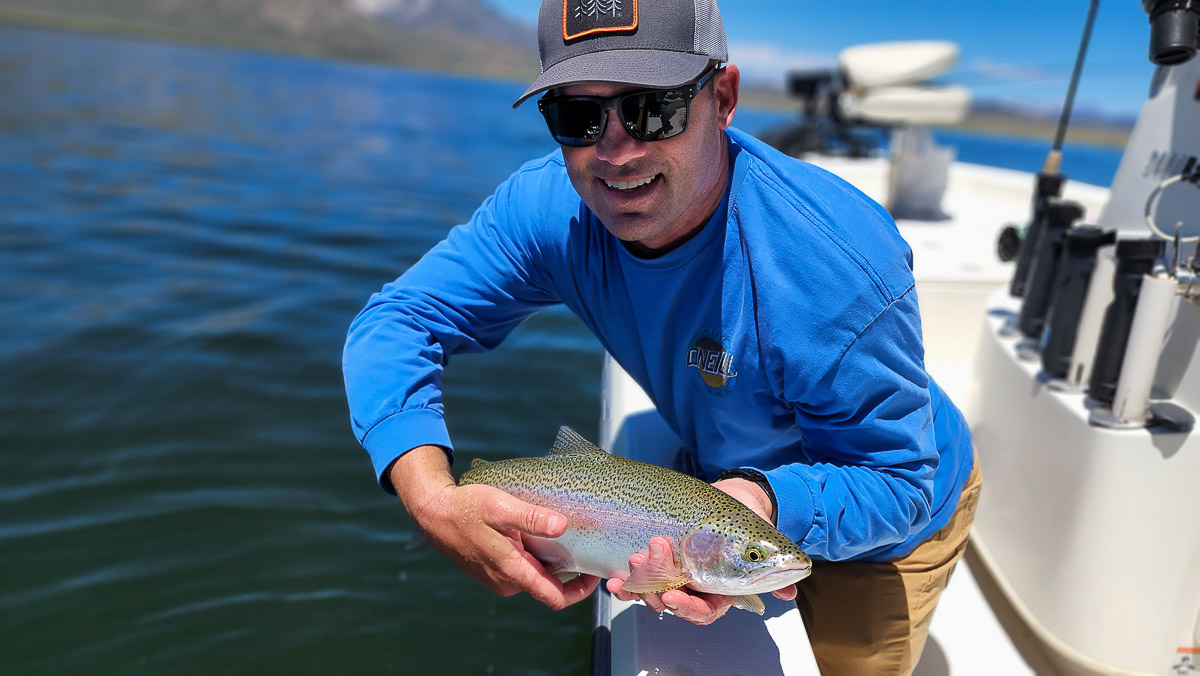 A smiling fly fisherman in a boat holding a large rainbow trout.