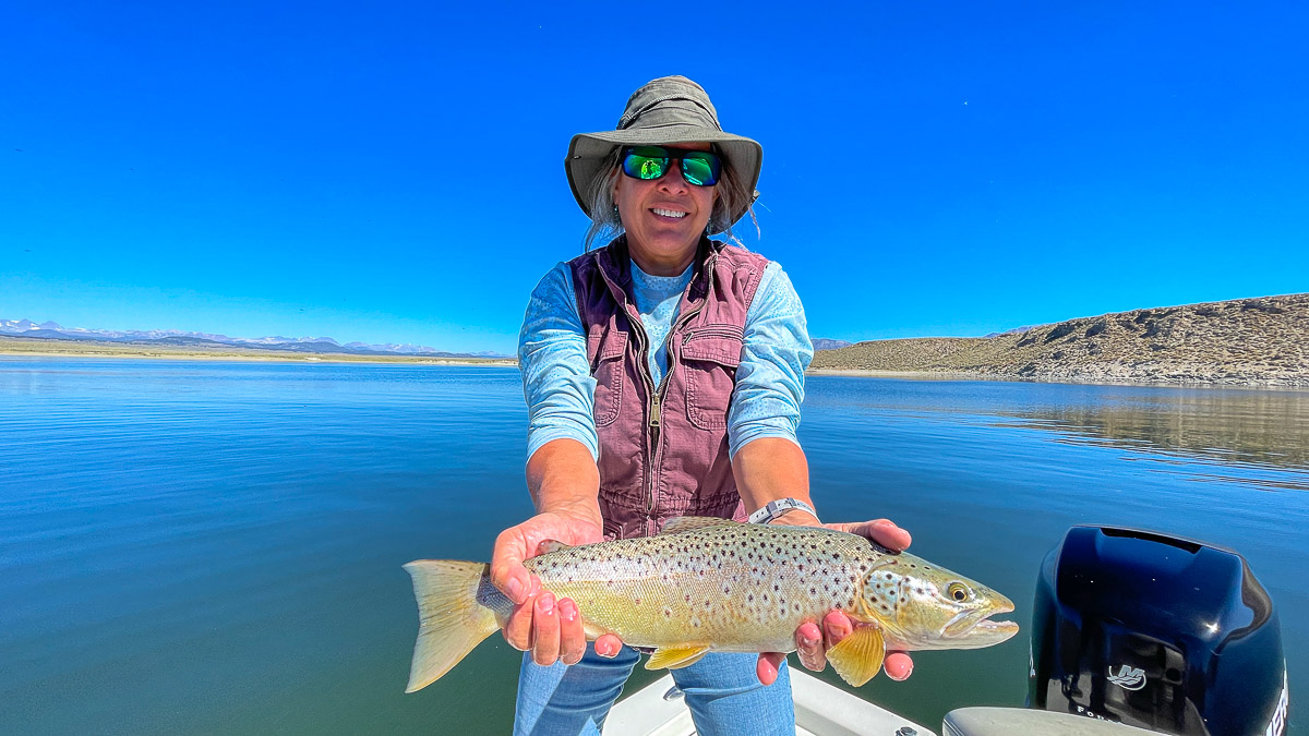 A fly fisherwoman on a lake in a boat holding a behemoth brown trout.