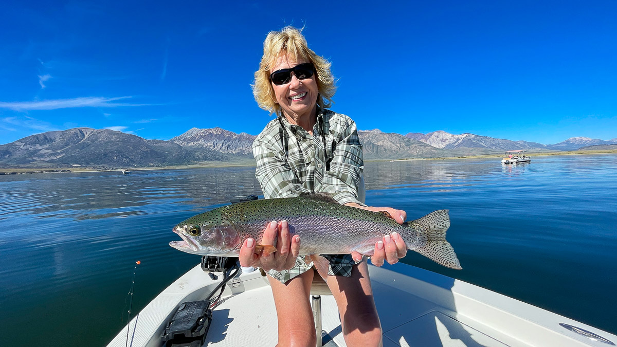 A smiling fly fisherwoman on a lake in a boat holding a large rainbow trout.