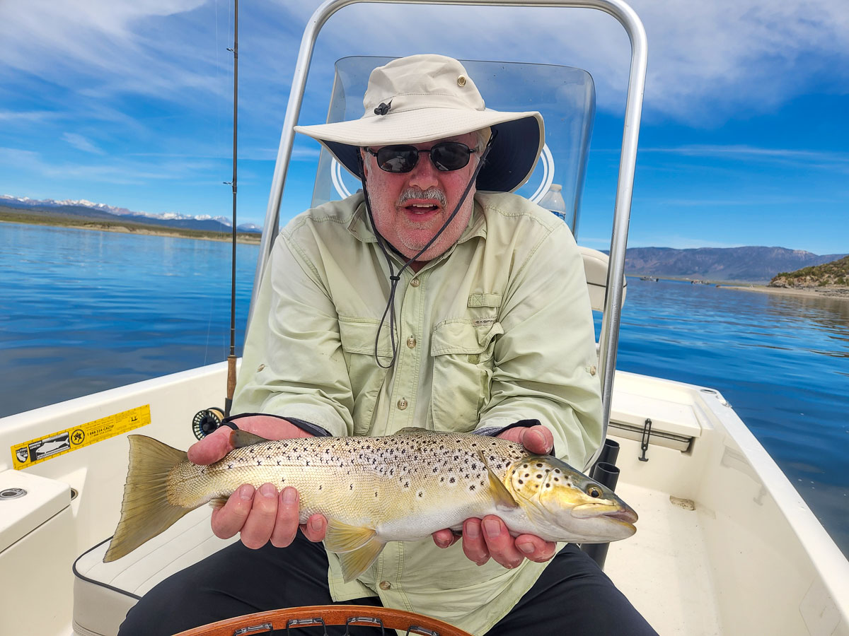 A fly fisherman on a lake in a boat holding a large brown trout.
