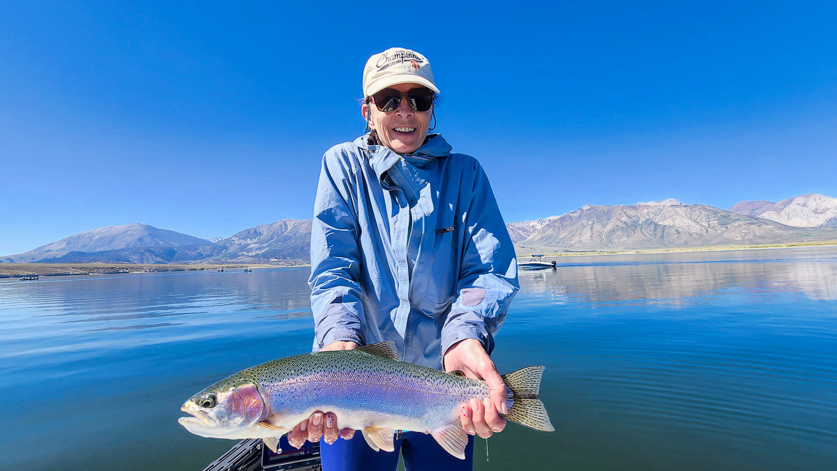 A fly fishermwoman on a lake in a boat holding a large rainbow trout.