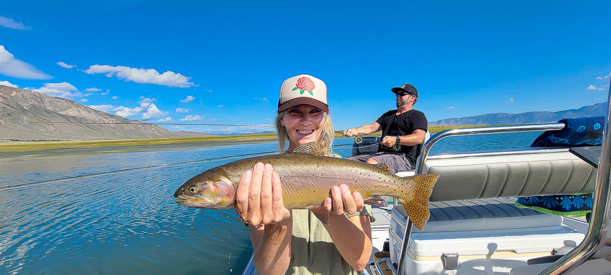 A fly fisherwoman on a lake in a boat holding a large cutthroat trout.
