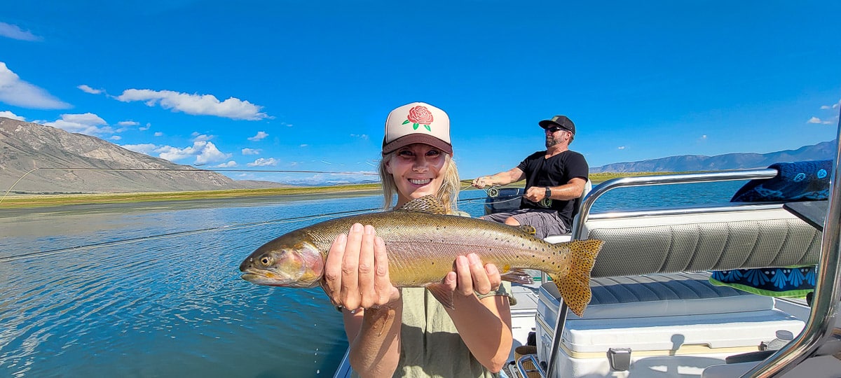 A fisherman holding a massive rainbow trout in a boat on a lake.