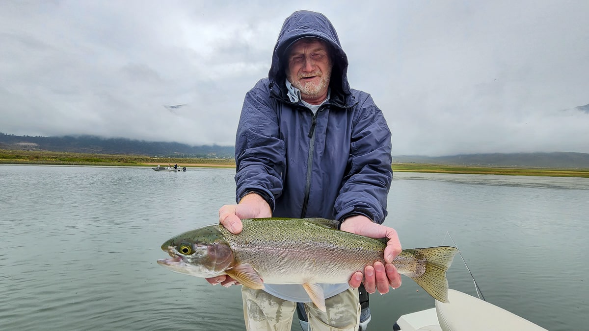 A fly fisherman on a lake in a boat holding a behemoth rainbow trout.