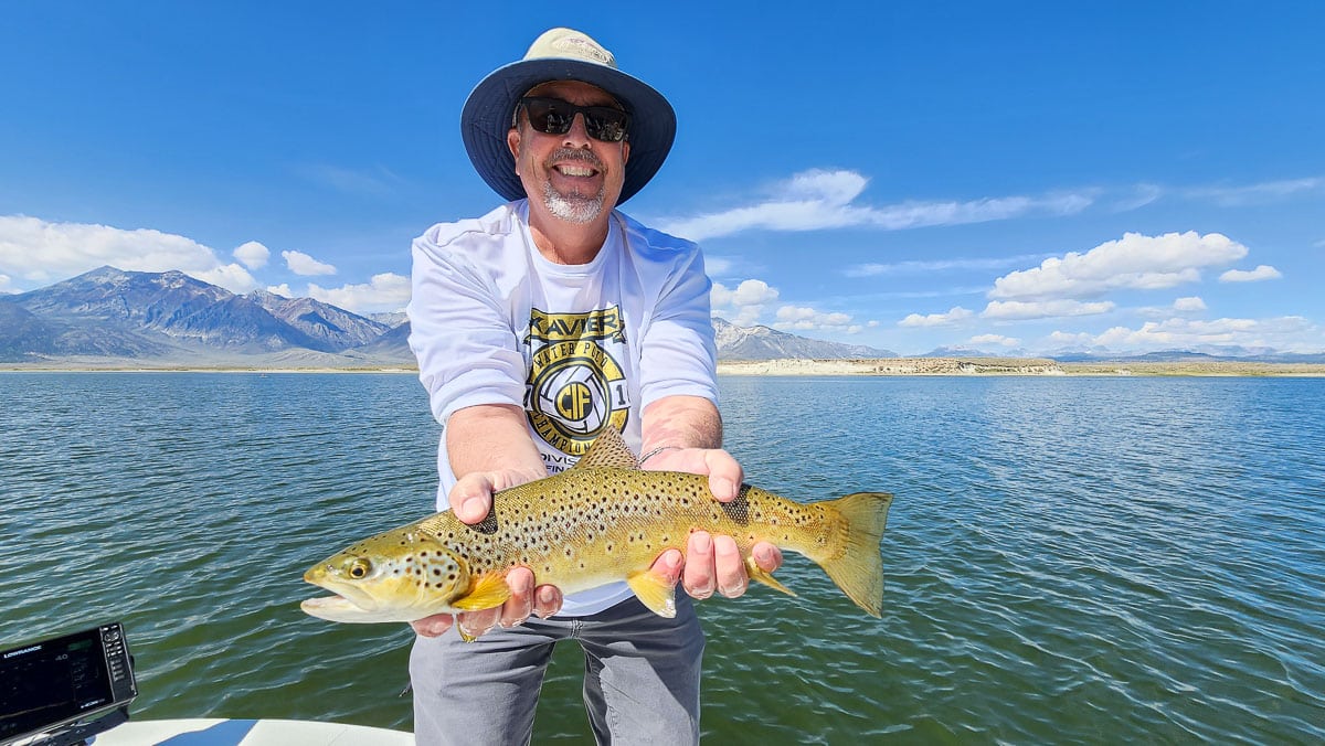 A smiling fly fisherman on a lake in a boat holding a large brown trout.