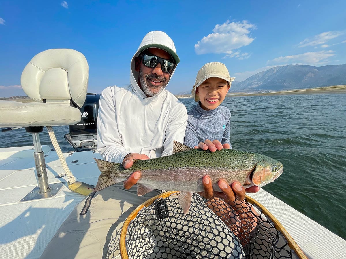 A smiling angler holding a large rainbow trout in a boat on a lake with a young boy.