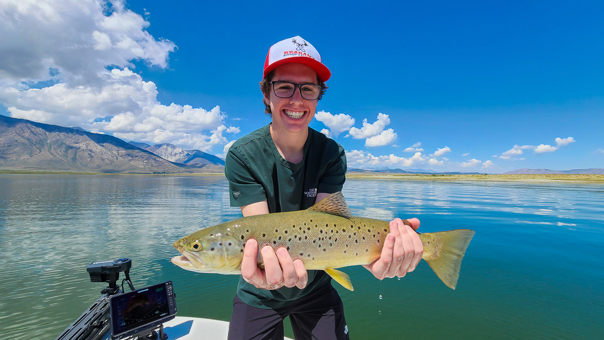 A smiling fly fisherman in a boat holding a large brown trout.