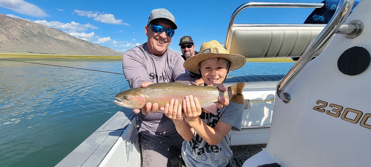 A smiling fly fisherman on a lake in a boat holding a large cutthroat trout next to a couple of kids.