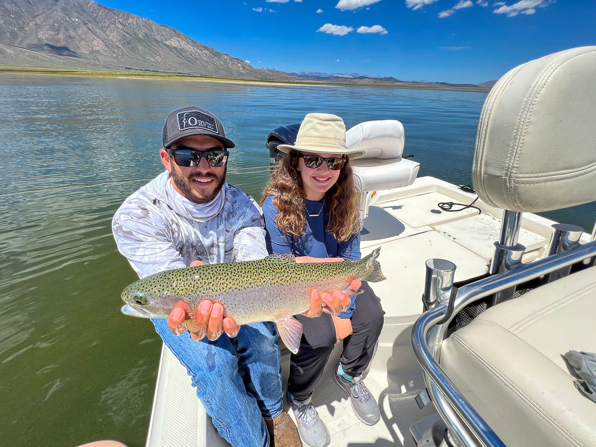 A smiling fly fisherman with a woman on a lake in a boat holding a large rainbow trout.