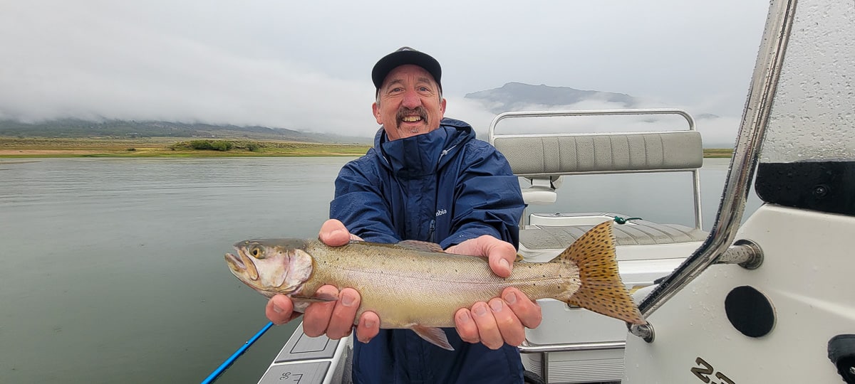 A smiling fly fisherman on a lake in a boat holding a large cutthroat trout.