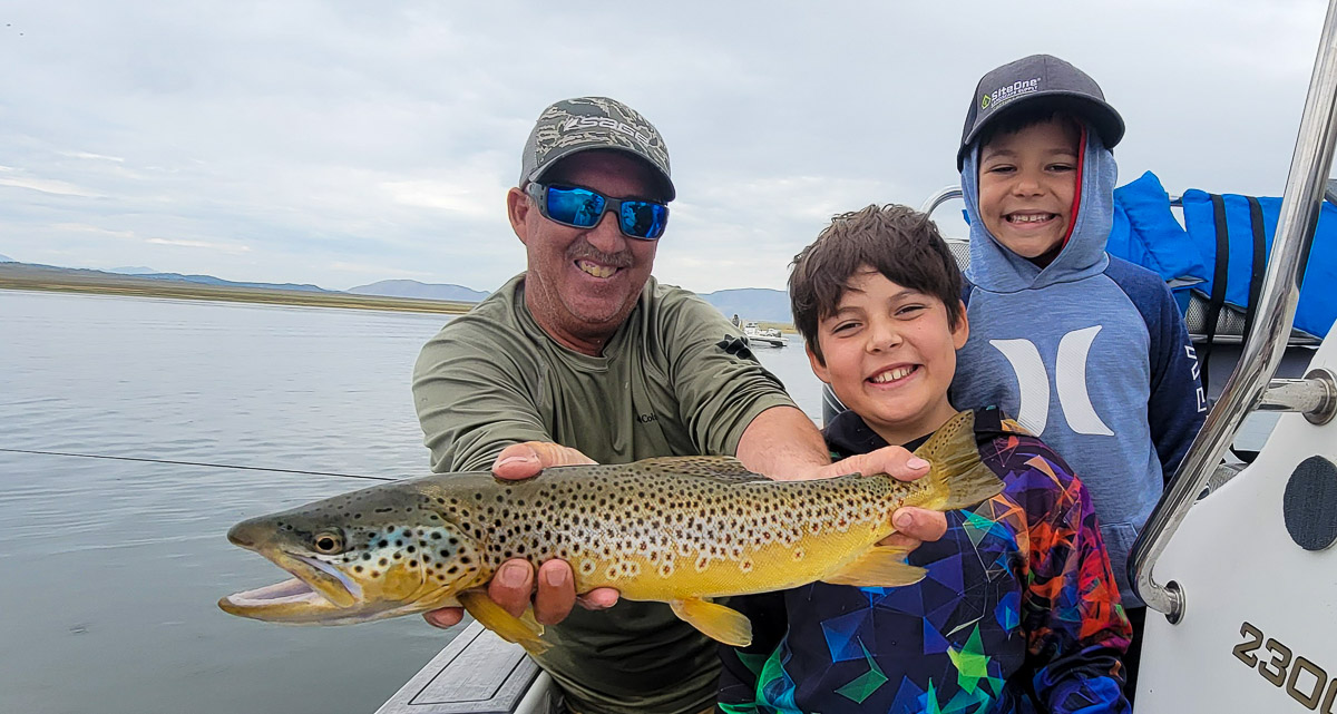 A fly fisherman on a lake in a boat holding a large brown trout and rainbow trout with a kid.