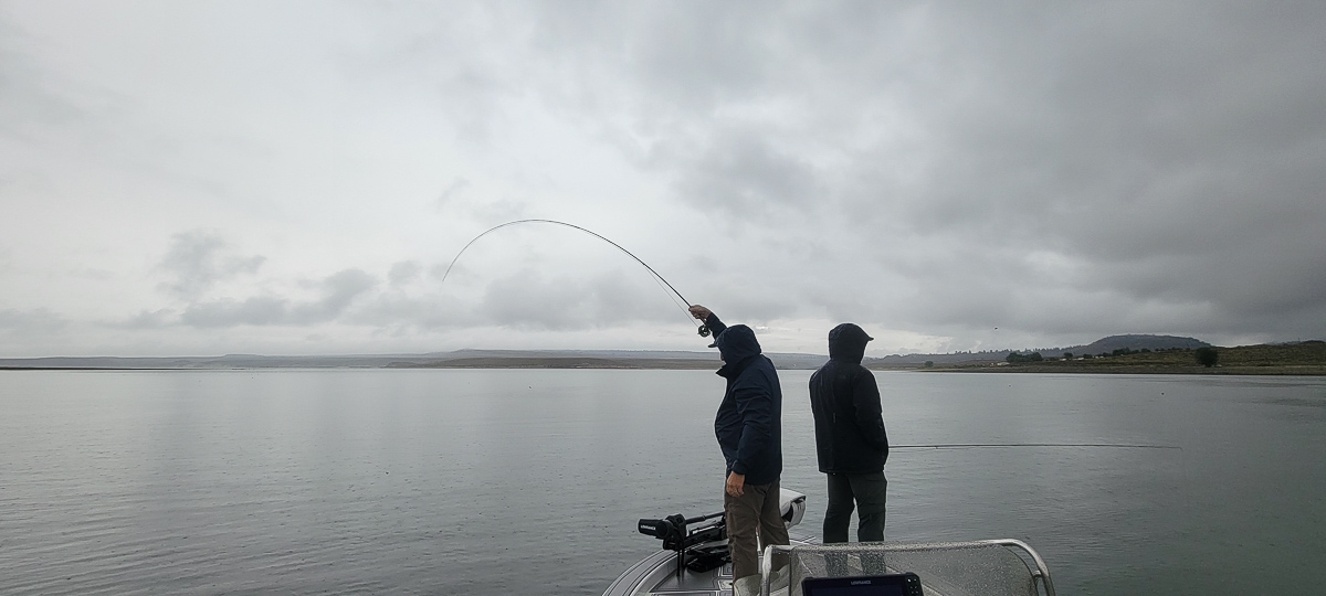 A fisherman with a bent rod hooked up to a fish on a lake on an overcast and rainy day.