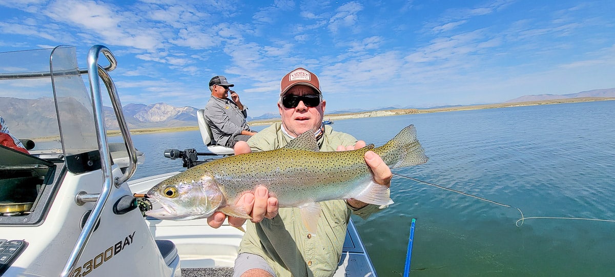 A smiling fly fisherman on a lake in a boat holding a large rainbow trout.