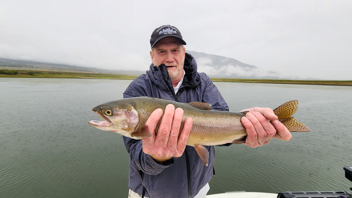 A smiling fly fisherman on a lake in a boat holding a large cutthroat trout.