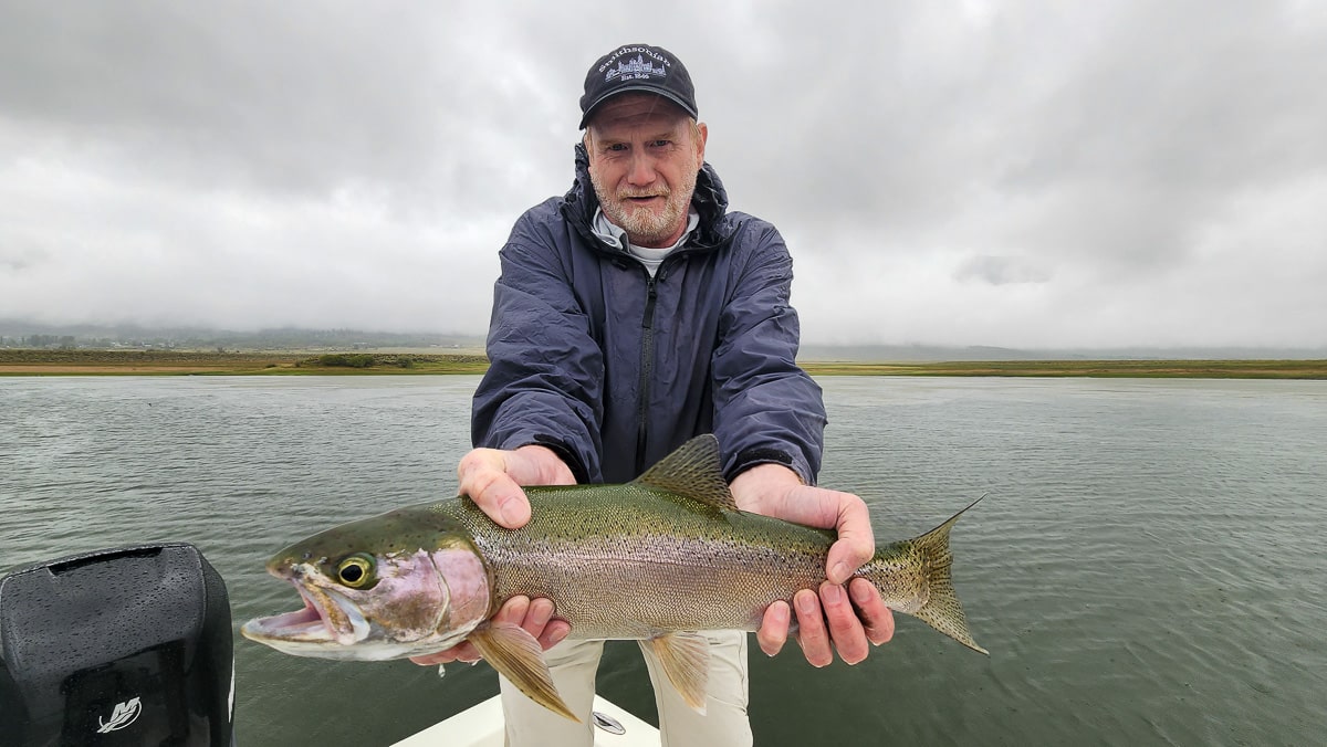 A fly fishermwoman on a lake in a boat holding a large rainbow trout.