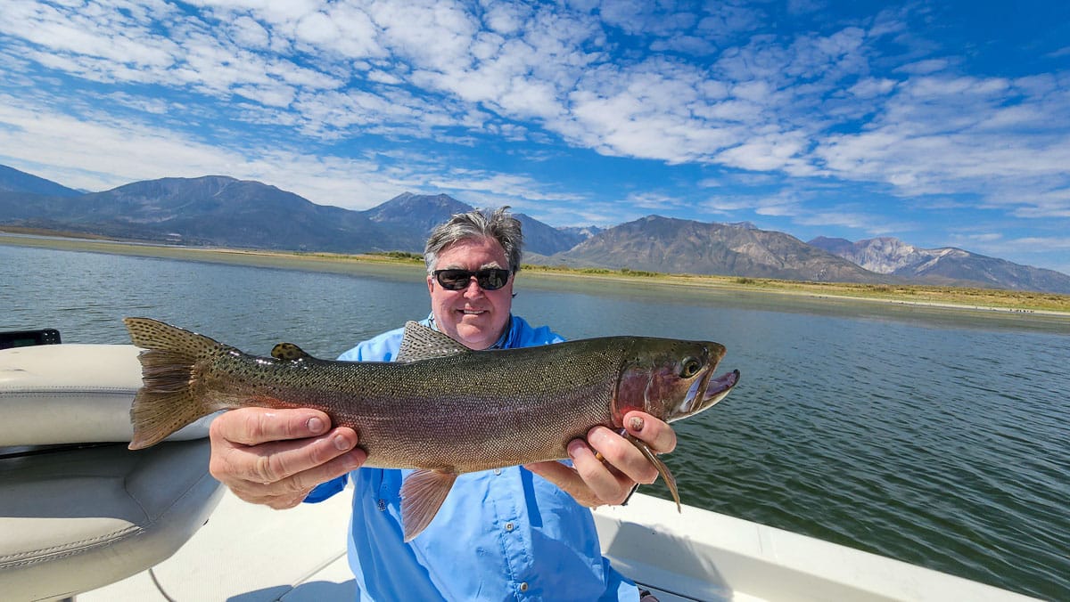 A fly fisherman on a lake in a boat holding a behemoth rainbow trout.