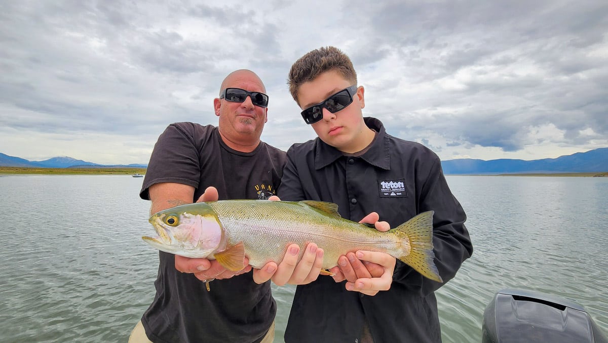 A pair of smiling anglers holding a large rainbow trout in a boat on a lake.