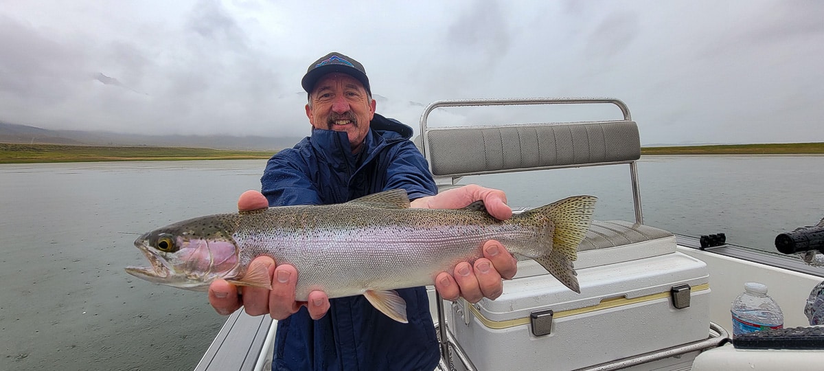 A smiling fly fisherman on a lake in a boat holding a large rainbow trout.