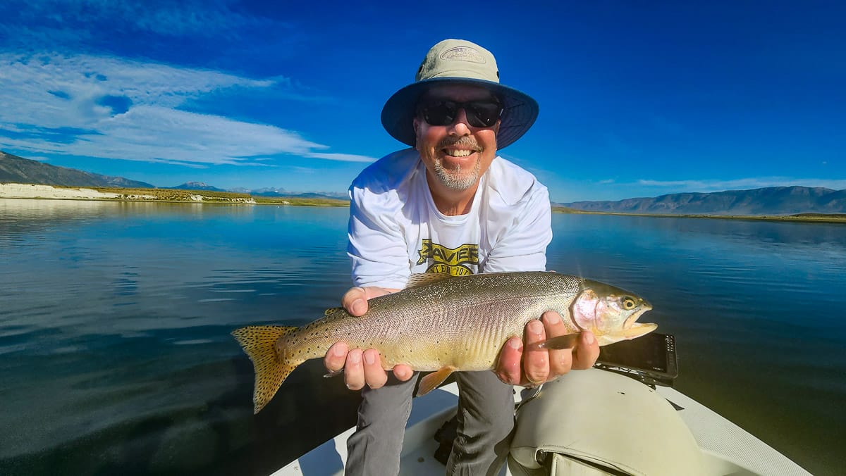 A fly fisherman on a lake in a boat holding a behemoth cutthroat trout.