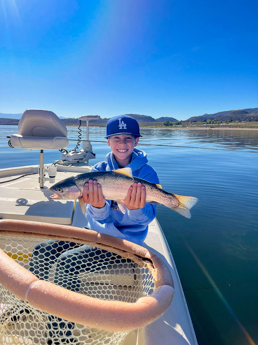 A smiling young fly fisherman holding a large rainbow trout in a boat with a net in the foreground.