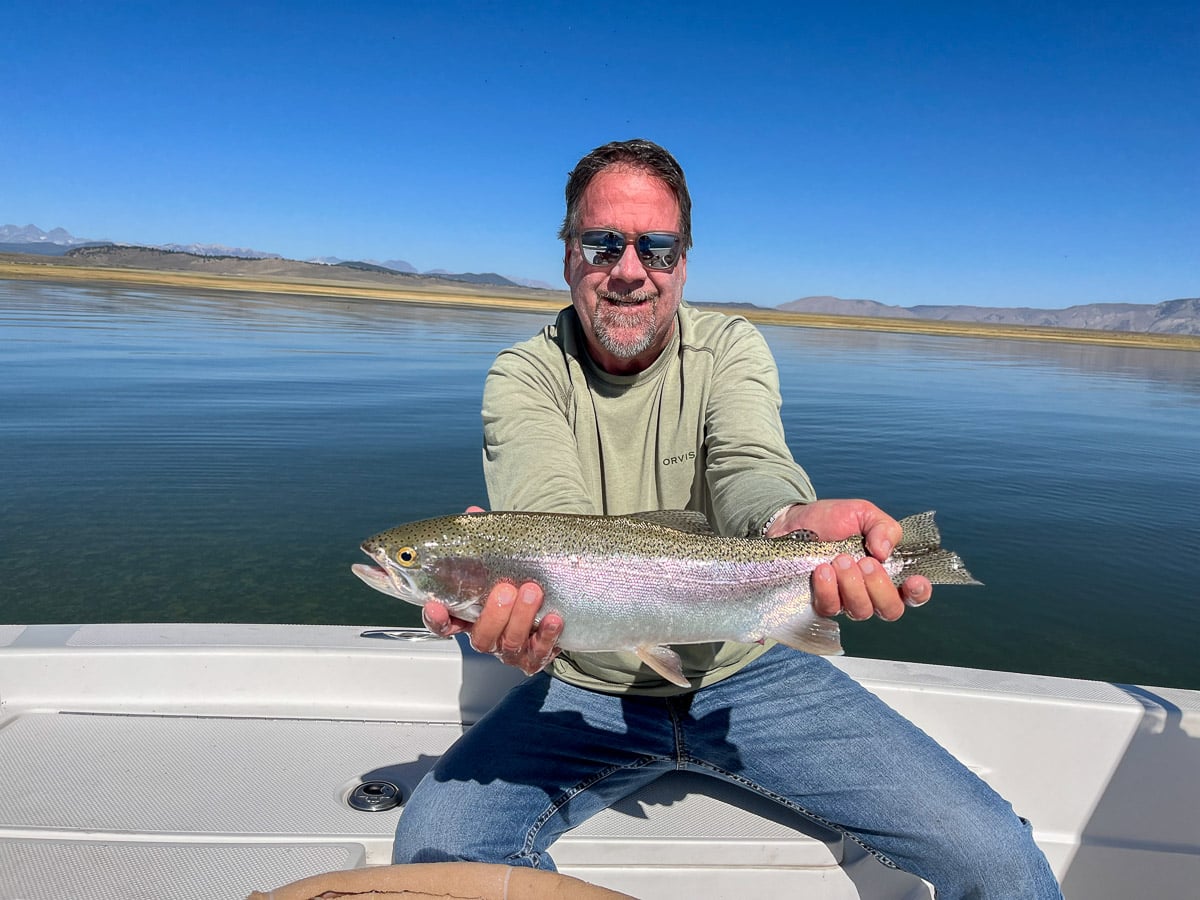 A smiling angler holding a large rainbow trout in a boat on a lake with a young boy.