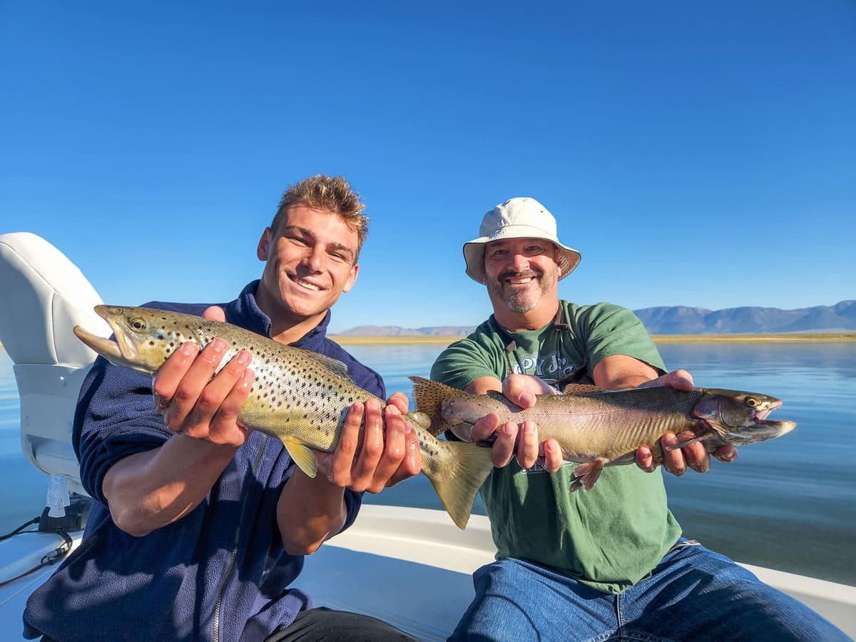 A smiling fly fisherman on a lake in a boat holding a large cutthroat trout.