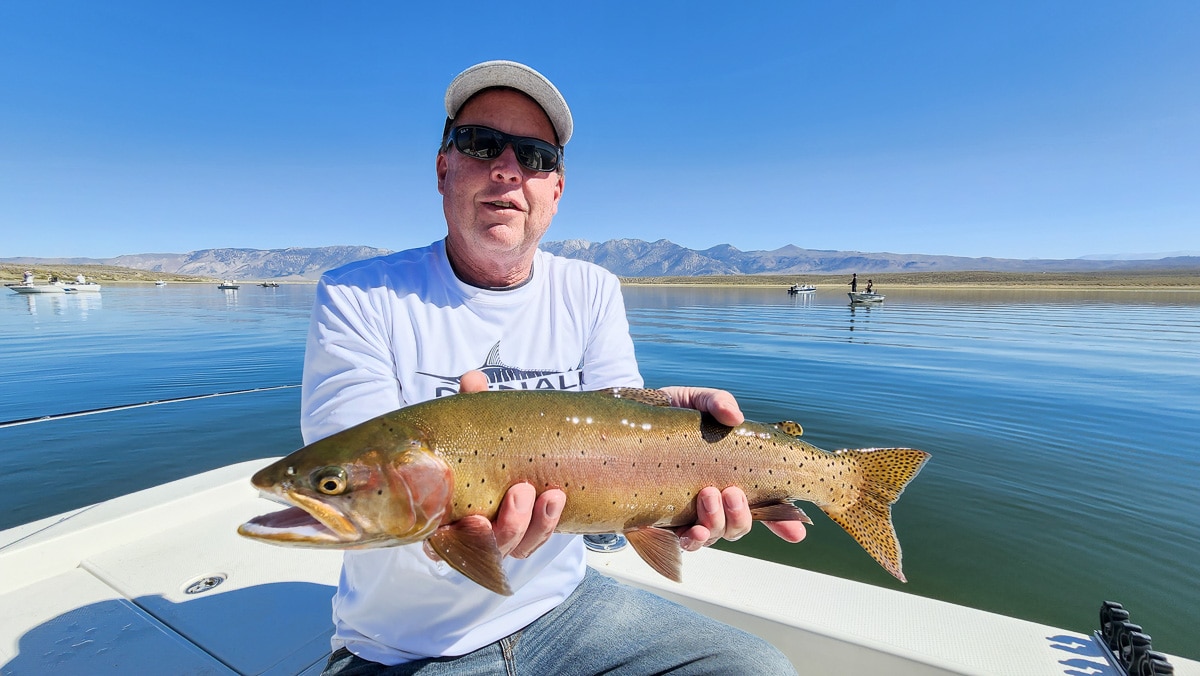 A smiling fly fisherman on a lake in a boat holding a large cutthroat trout.