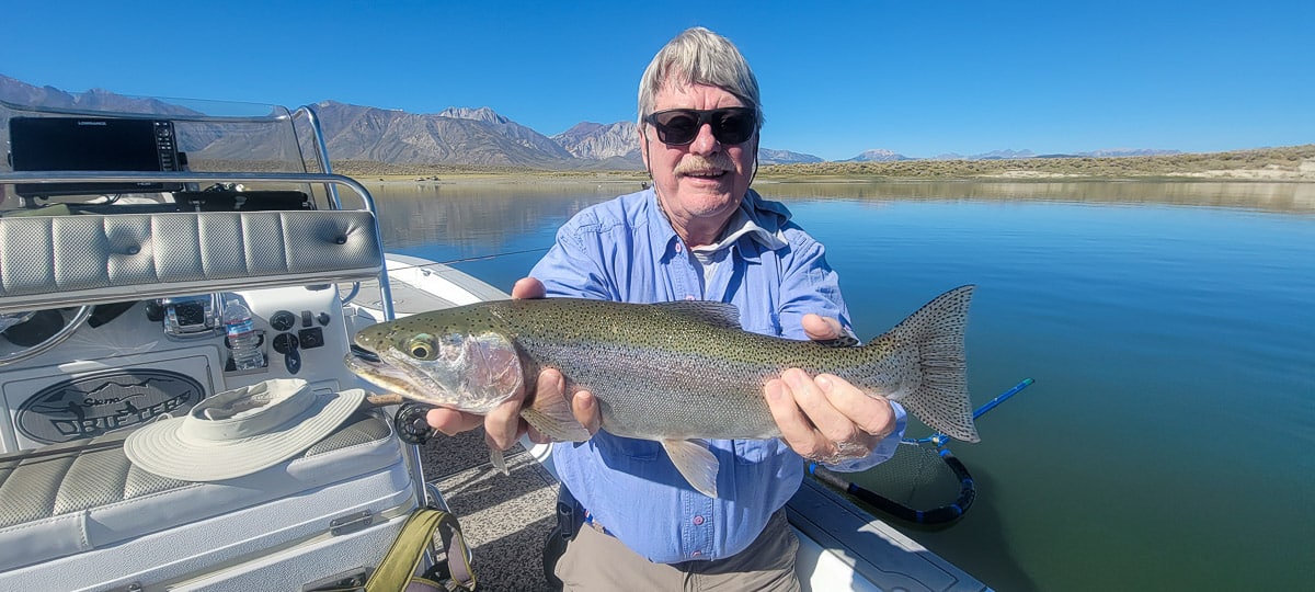 A smiling fly fisherman on a lake in a boat holding a large cutthroat trout.