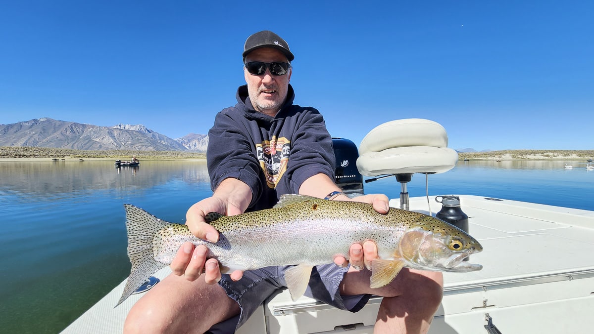 A fly fishermwoman on a lake in a boat holding a large rainbow trout.