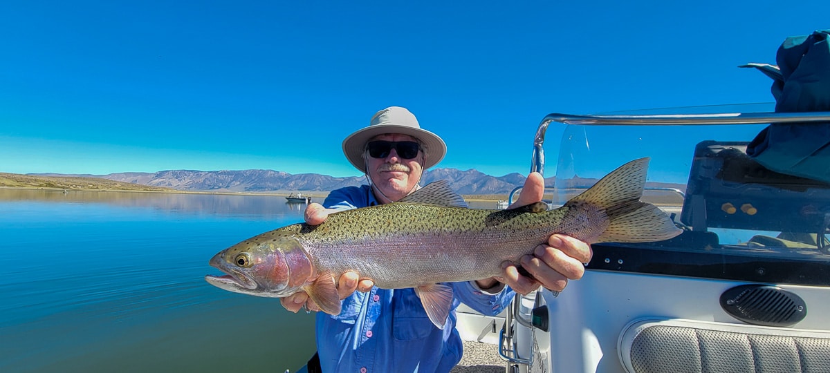 A smiling fly fisherman on a lake in a boat holding a large rainbow trout.