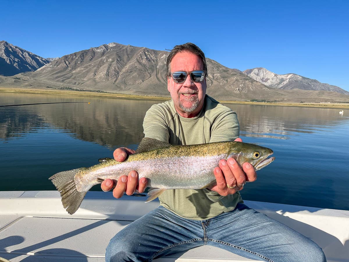 A fly fisherman on a lake in a boat holding a behemoth rainbow trout.