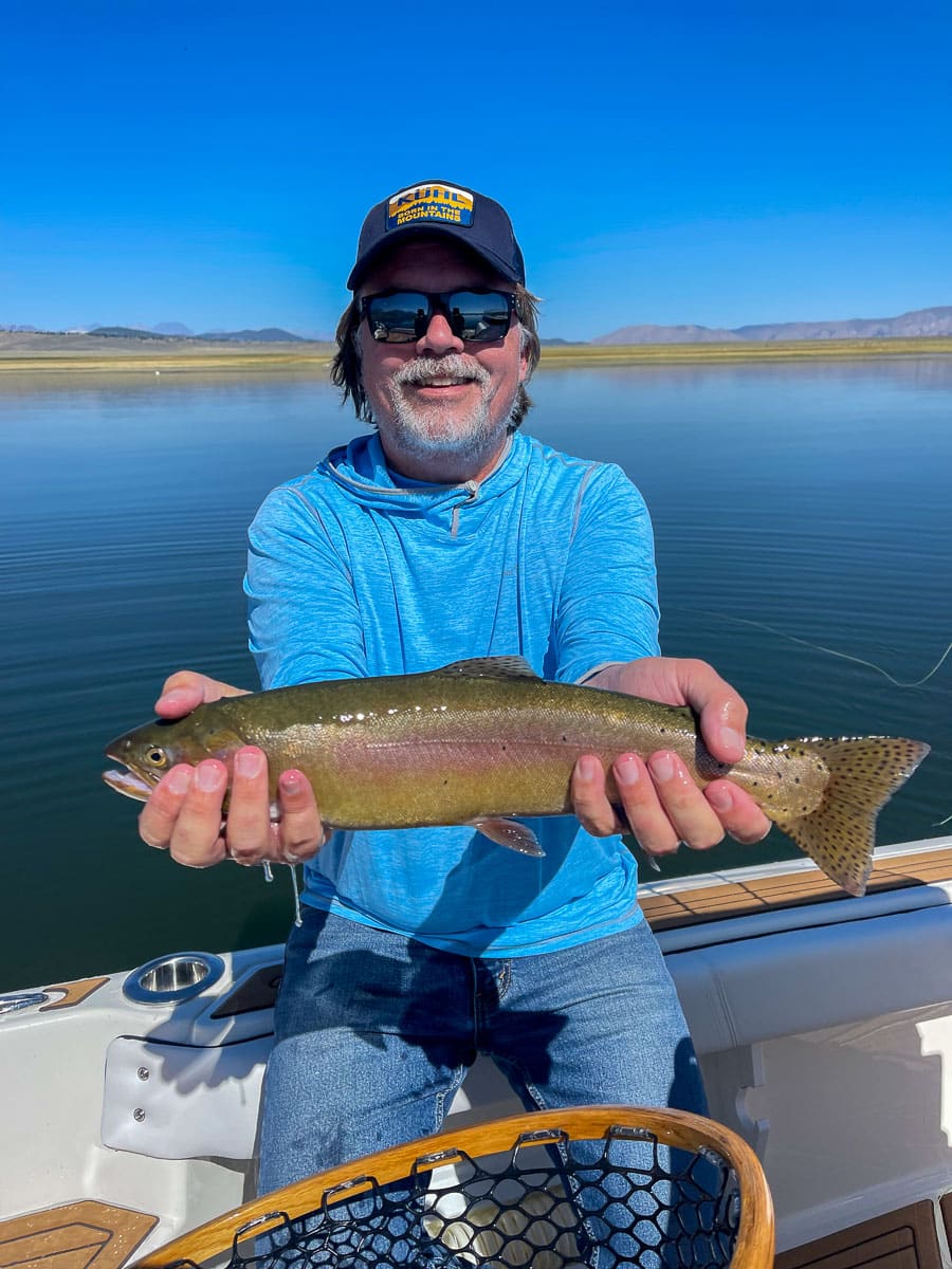 A smiling angler holding a large rainbow trout in a boat on a lake with a young boy.