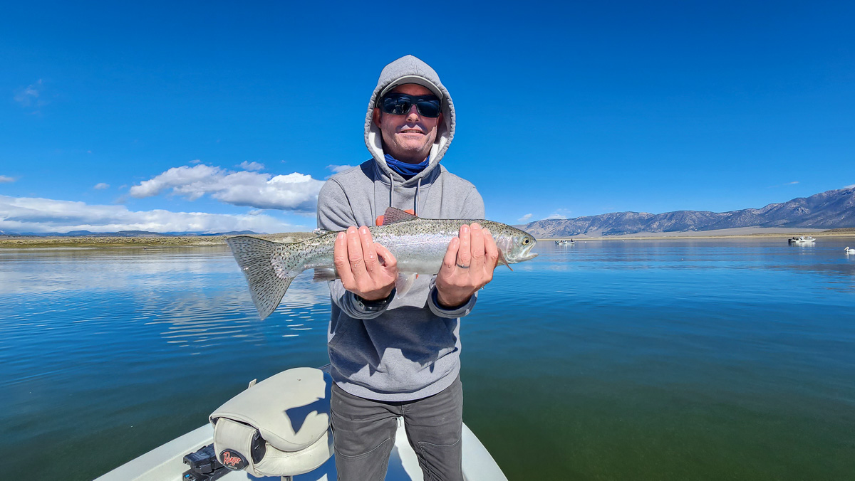 A fly fisherman on a lake in a boat holding a large rainbow trout.