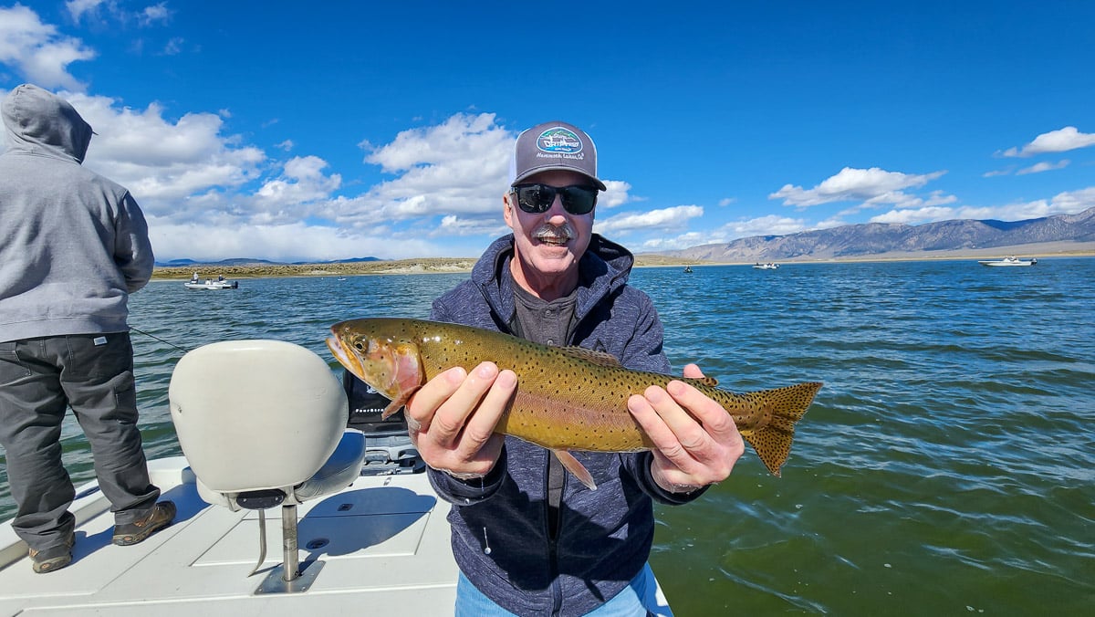 A smiling fly fisherman on a lake in a boat holding a large cutthroat trout.