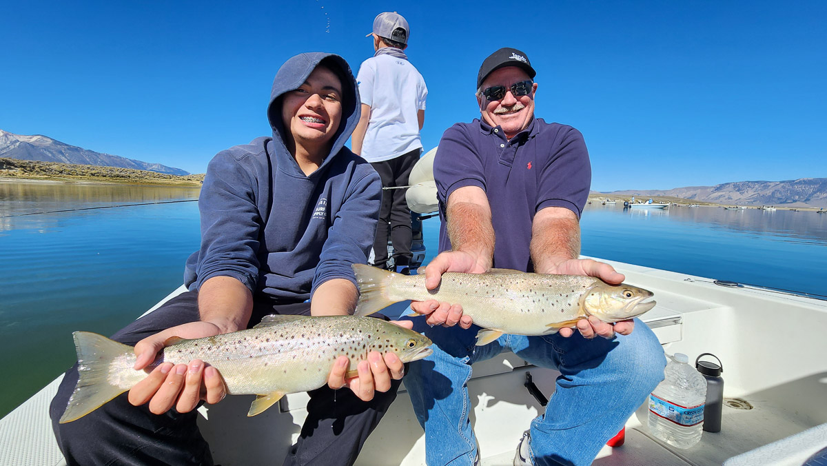 A pair of fishermen holding two large brown trout in a boat on a lake.