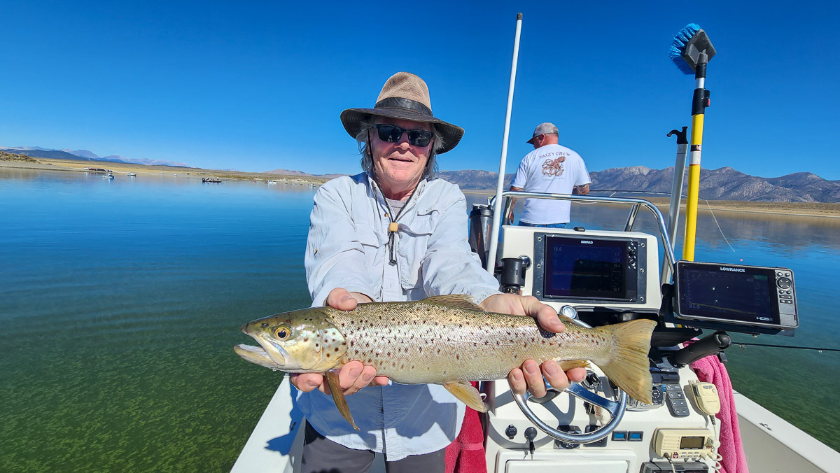 A smiling fly fisherman on a lake in a boat holding a large rainbow trout.