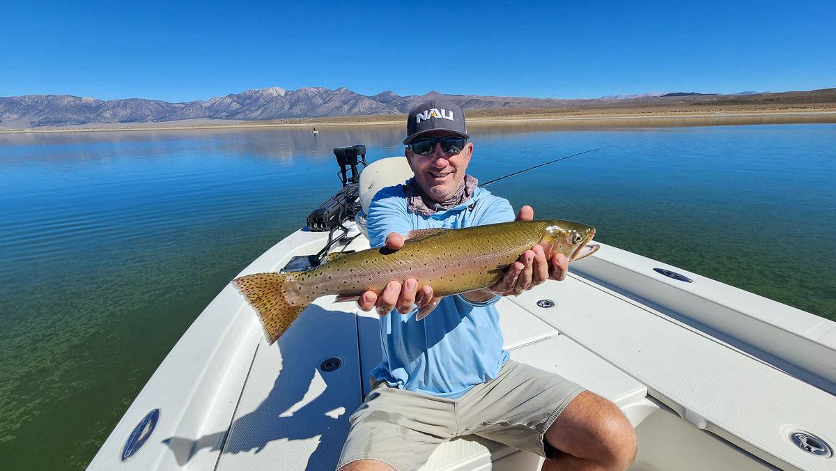 A smiling fly fisherman on a lake in a boat holding a large cutthroat trout.