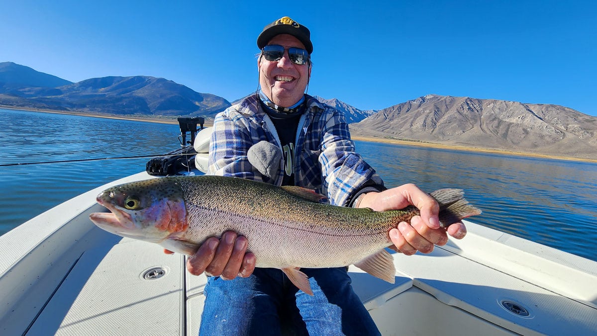 A smiling fly fisherman on a lake in a boat holding a large rainbow trout.