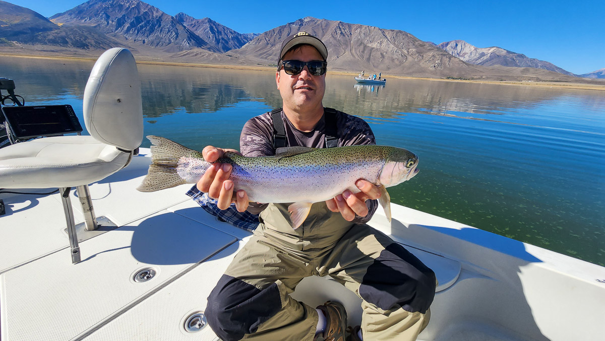 A smiling fly fisherman on a lake in a boat holding a large rainbow trout.