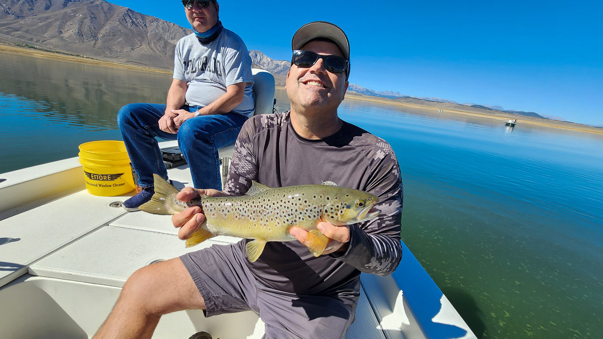 A smiling fly fisherman on a lake in a boat holding a large brown trout.