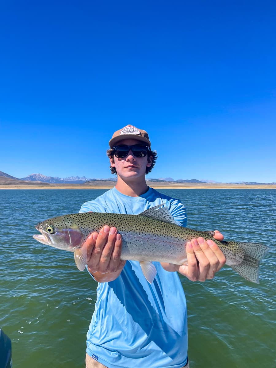 A fly fisherman on a lake in a boat holding a behemoth rainbow trout.