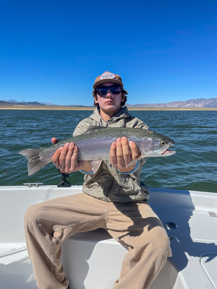 A smiling angler holding a large rainbow trout in a boat on a lake with a young boy.