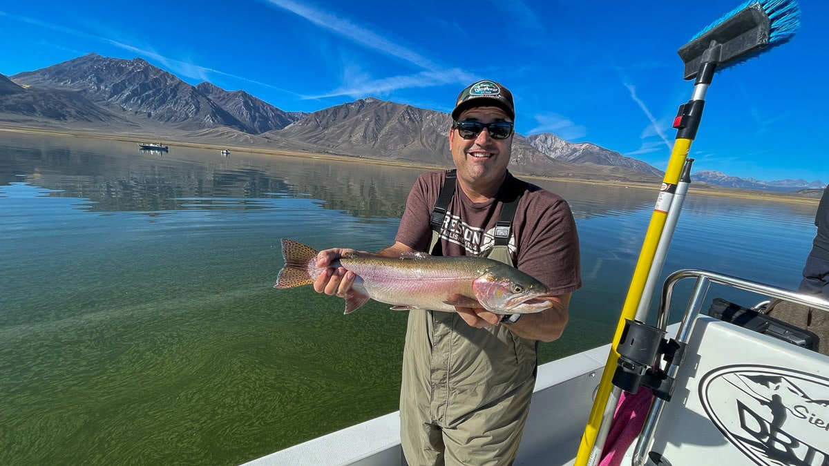 A smiling fly fisherman on a lake in a boat holding a large cutthroat trout.