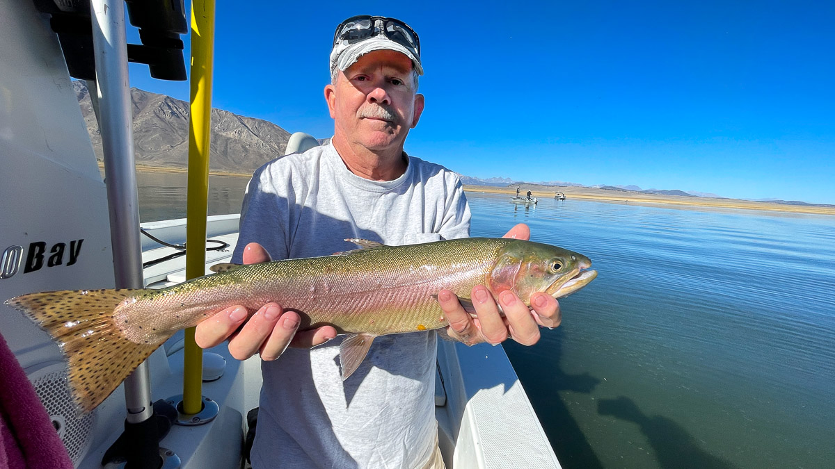A smiling fly fisherman on a lake in a boat holding a large cutthroat trout.