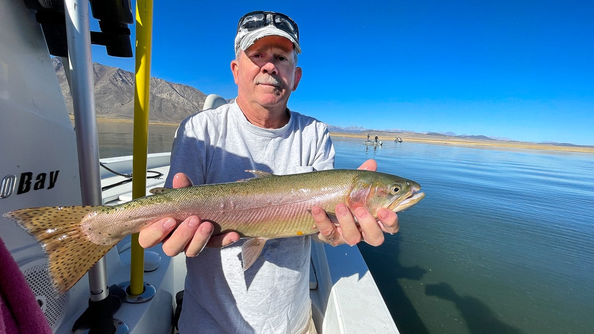 A smiling fly fisherman on a lake in a boat holding a large cutthroat trout.